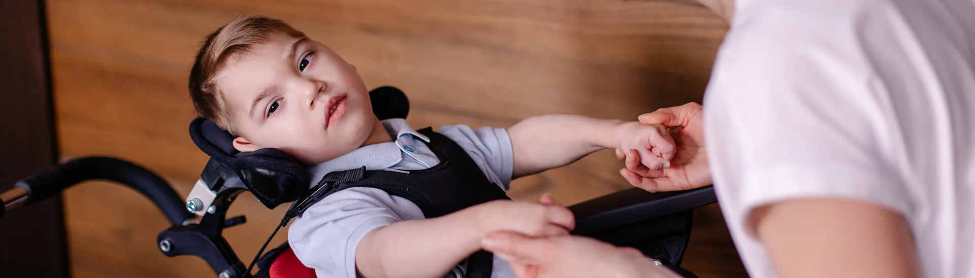 A handicap little boy with his head resting on a wheelchair head rest looking up while a woman holds his hand.
