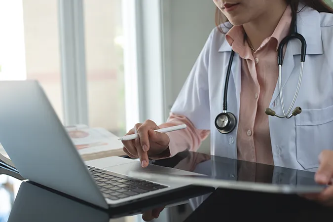 Female-Doctor-At-Computer_2470322883 A female doctor with a stethoscope around her neck is adding notes on her laptop.