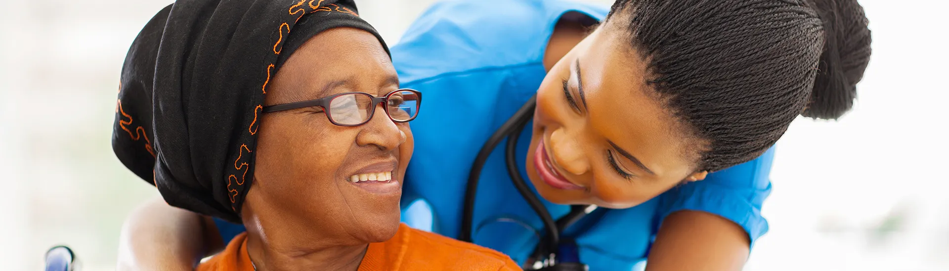 A younger nurse with her arms wrapped supportively around an elder woman's shoulders while she sits.