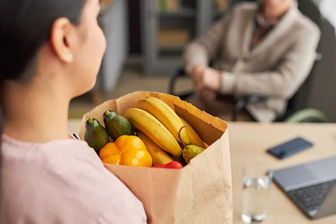 A woman holding a paper bag full of fruits and vegetables while talking to a woman in a wheelchair.